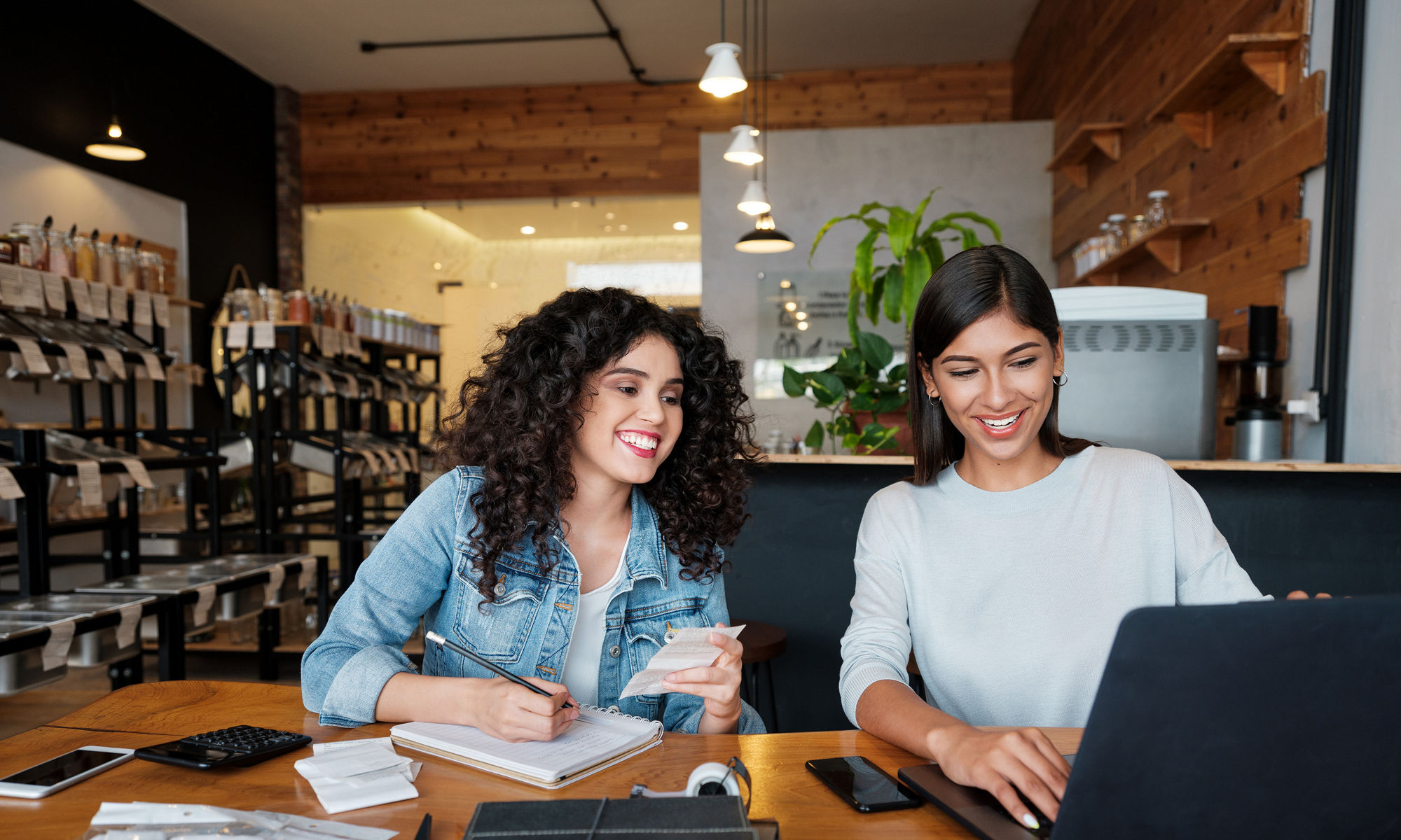 Two young latin women sitting at a table in an eco-friendly store, using a laptop and smiling.