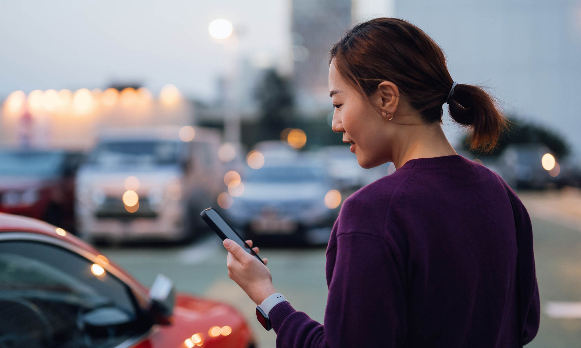 Young Asian businesswoman using smartphone while walking to her car, commuting in the city, with urban cityscape in background. Business on the go concept